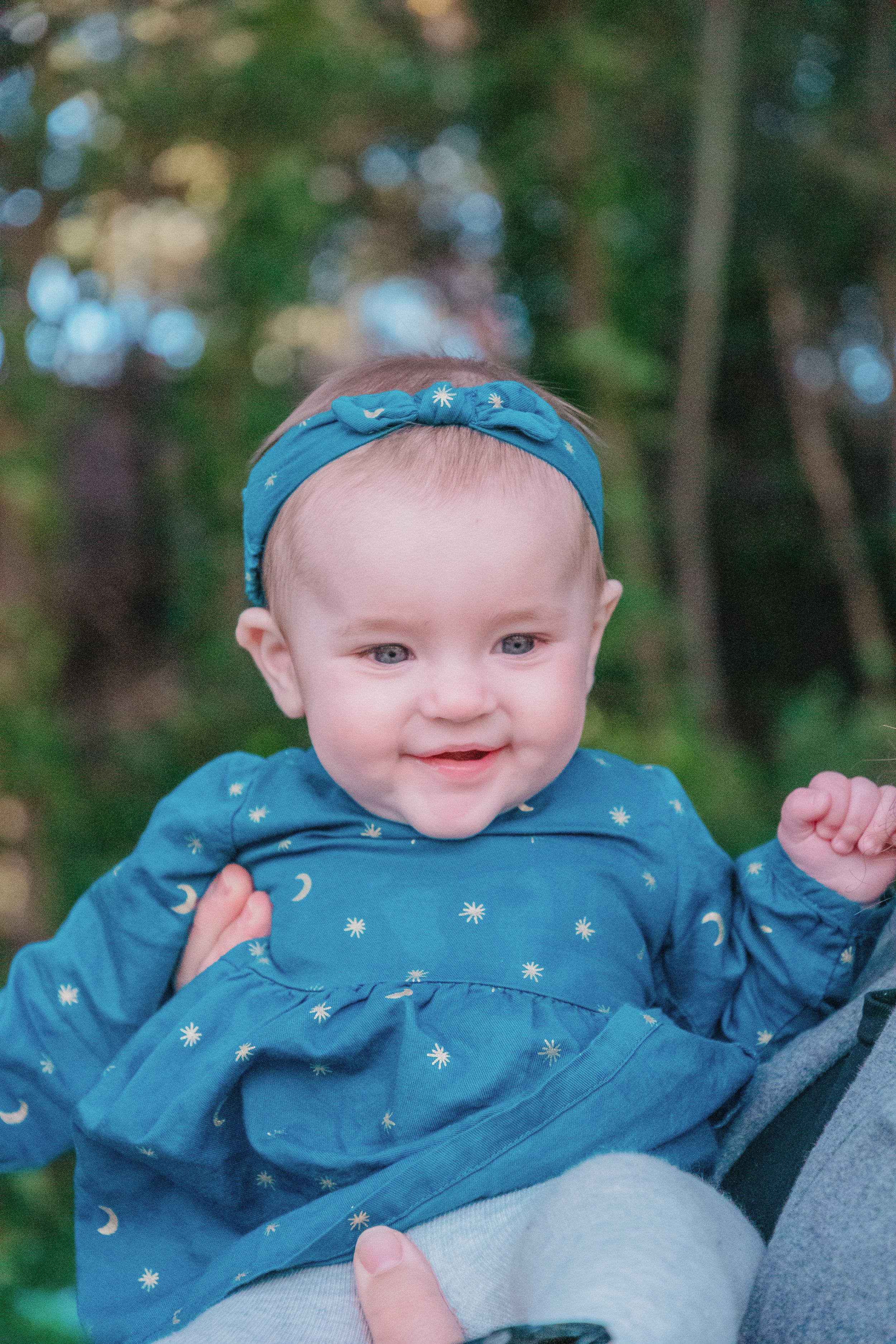 Baby wearing blue patterned dress and matching headband smiling outdoors.
