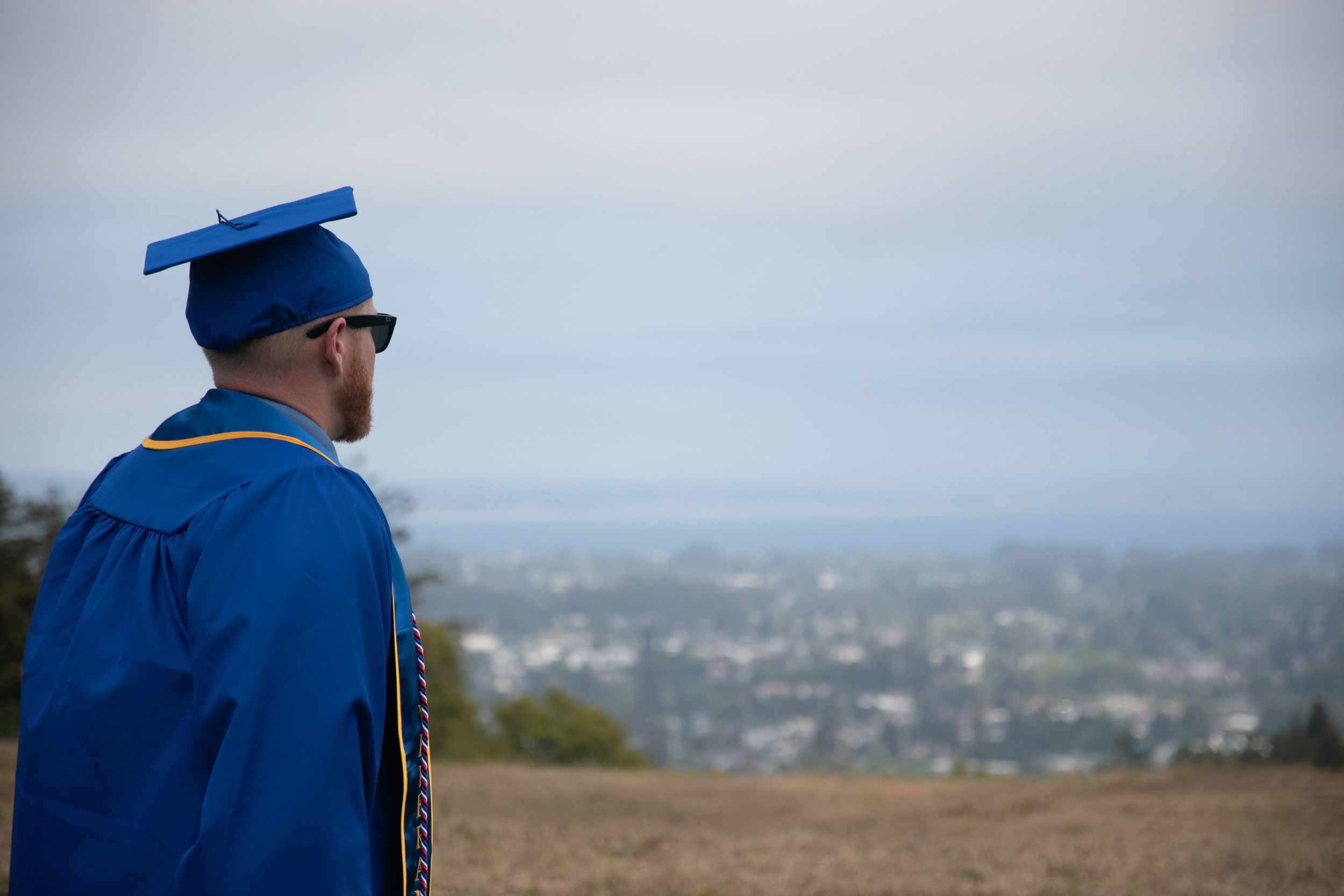 A graduate in blue cap and gown looks out over a city vista on graduation day.