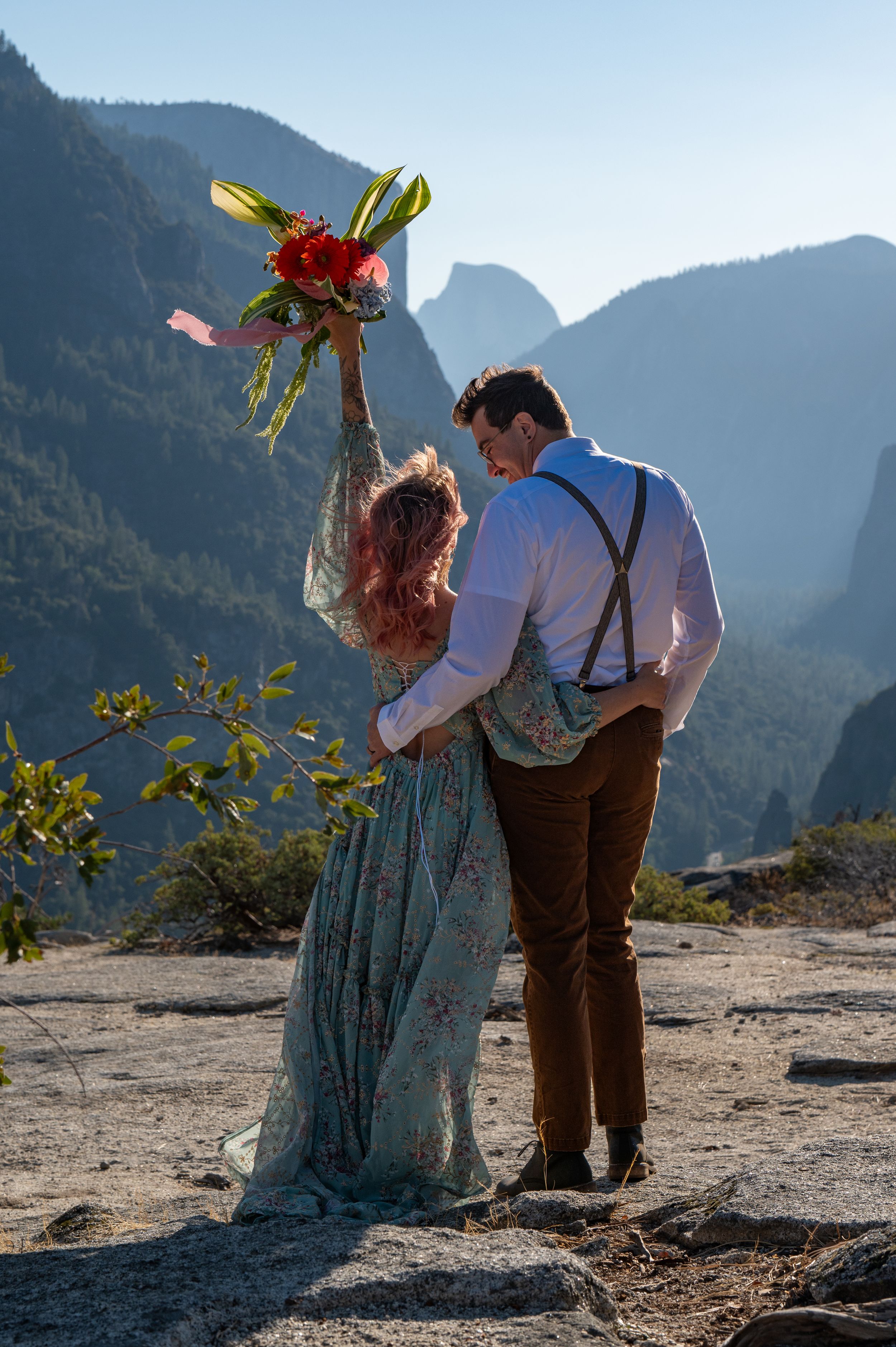 A couple celebrates their engagement with a bouquet held high against a mountainous backdrop.