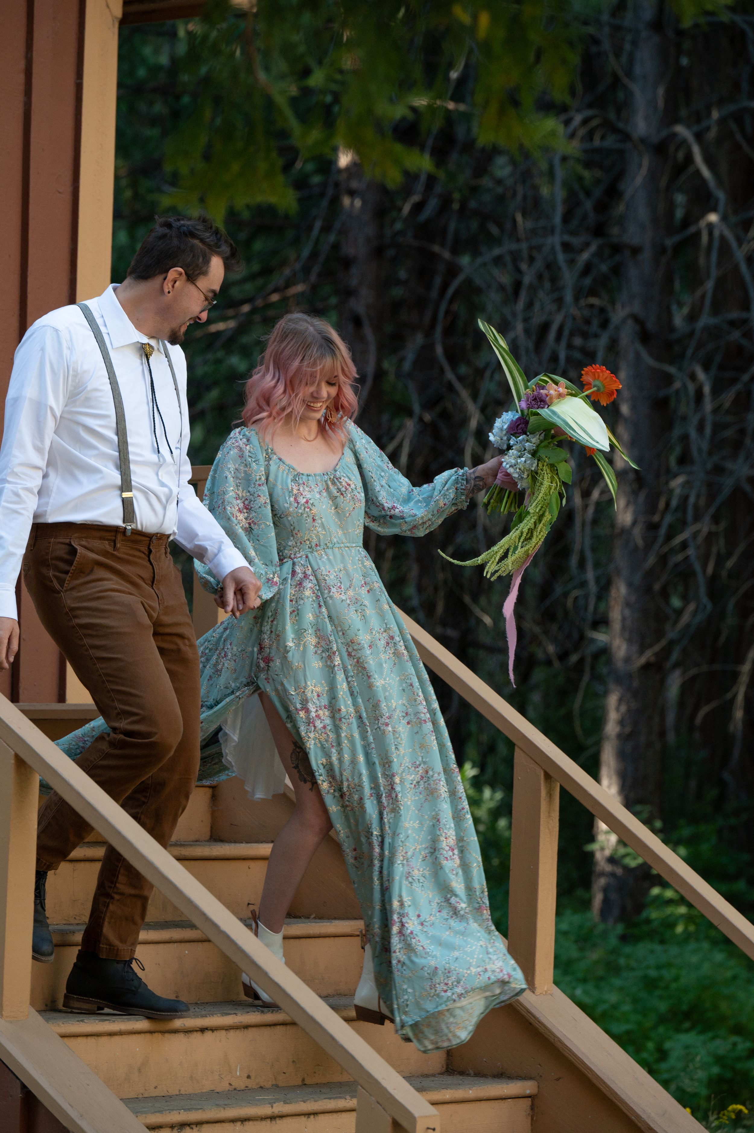 A couple descends wooden stairs, with the person in a flowing turquoise dress holding a bouquet.