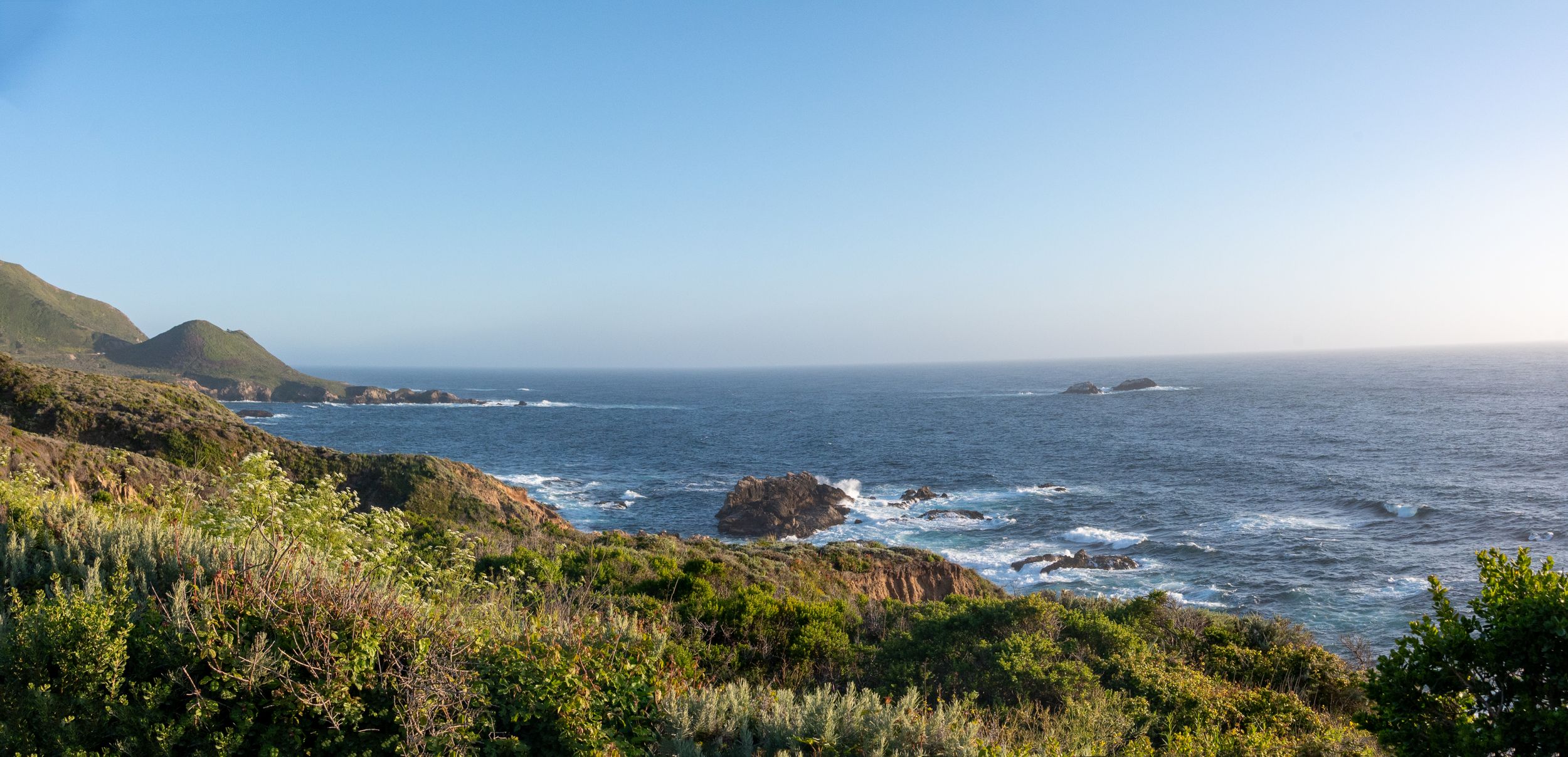 Panoramic coastal landscape showing rocky cliffs and blue ocean waves against a clear sky.