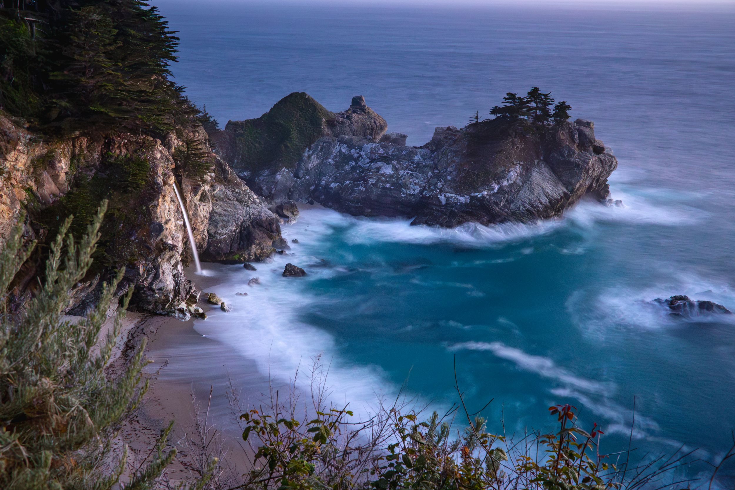 Dramatic coastal view of McWay Falls with turquoise waters crashing against rocky cliffs in Big Sur, California.