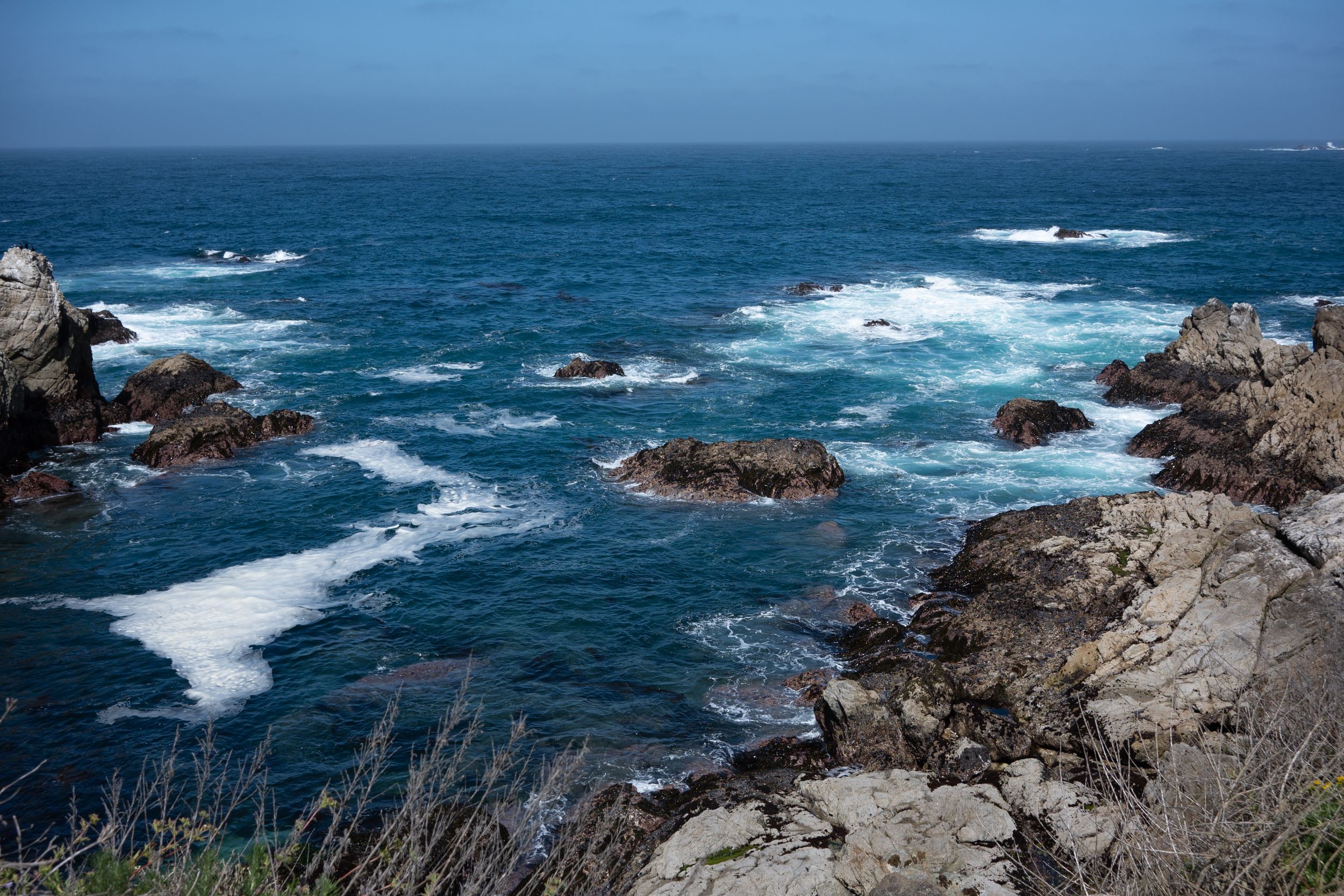 Rugged coastline with white waves crashing against rocky outcrops in the deep blue ocean.