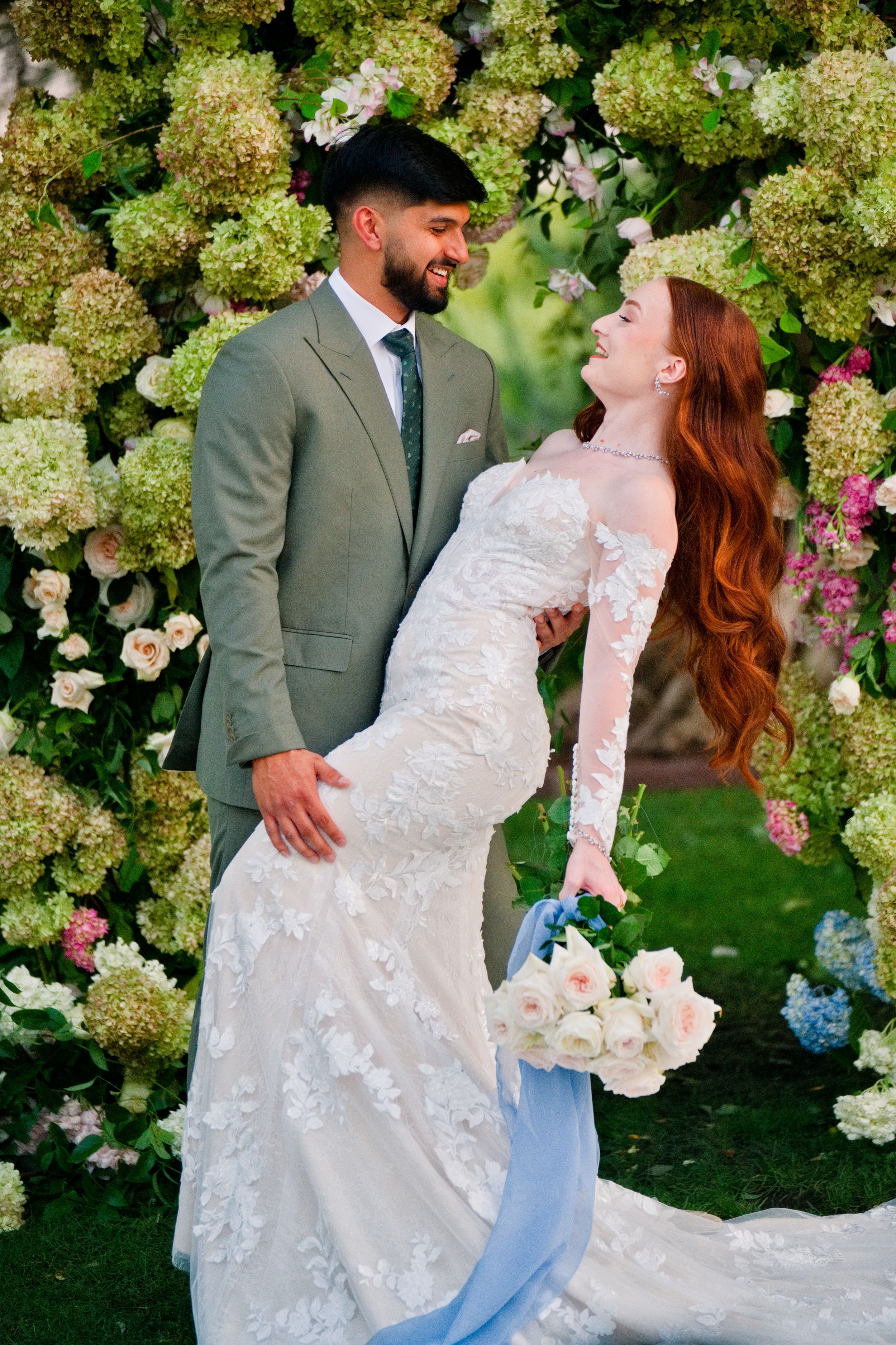 Newlyweds pose against a lush wall of hydrangeas and flowers in a romantic garden setting.