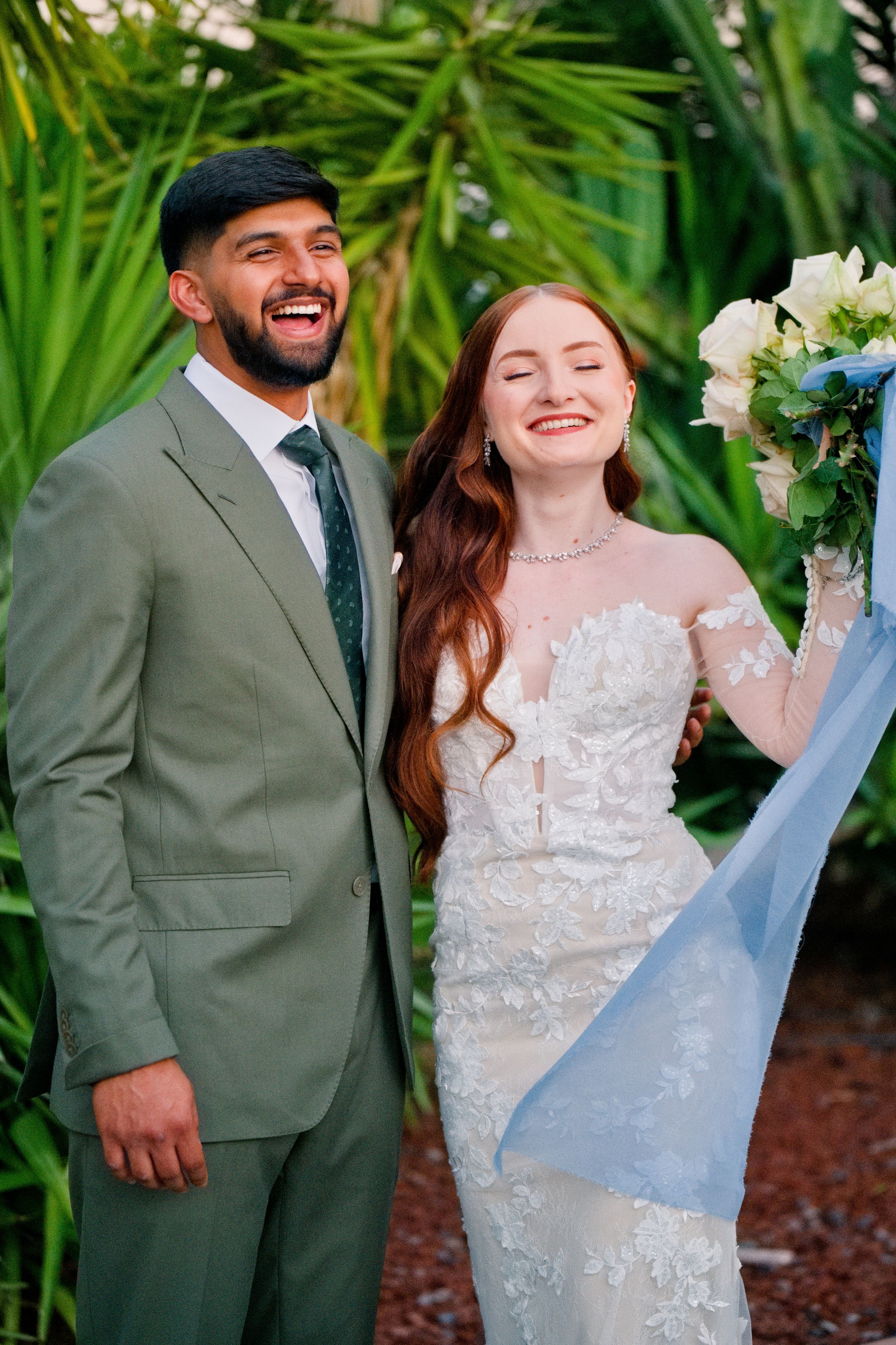 A joyful wedding couple laughs together against a backdrop of tropical palms.