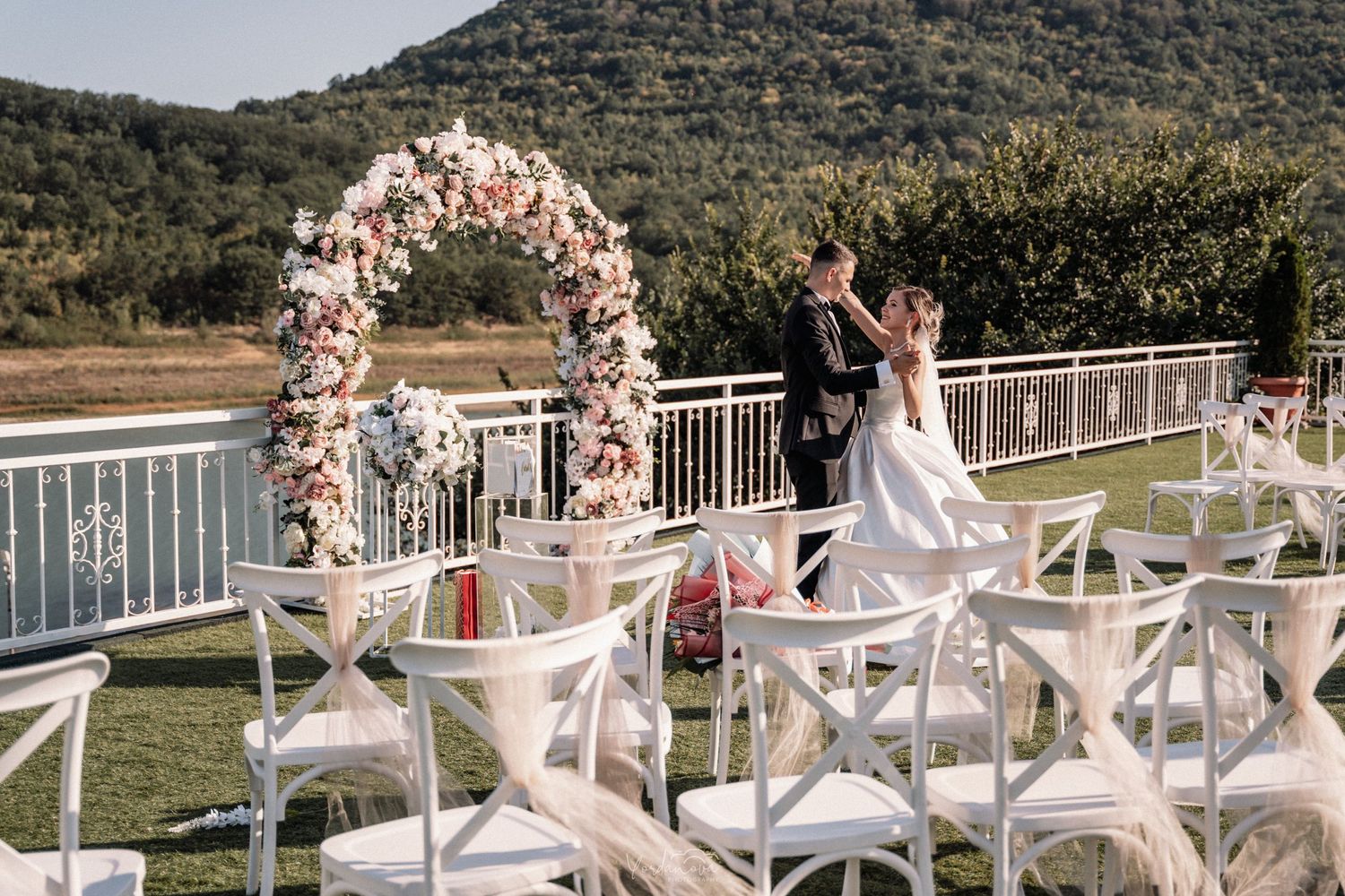Elegant outdoor wedding ceremony setup with floral arch and white chairs against mountain backdrop.