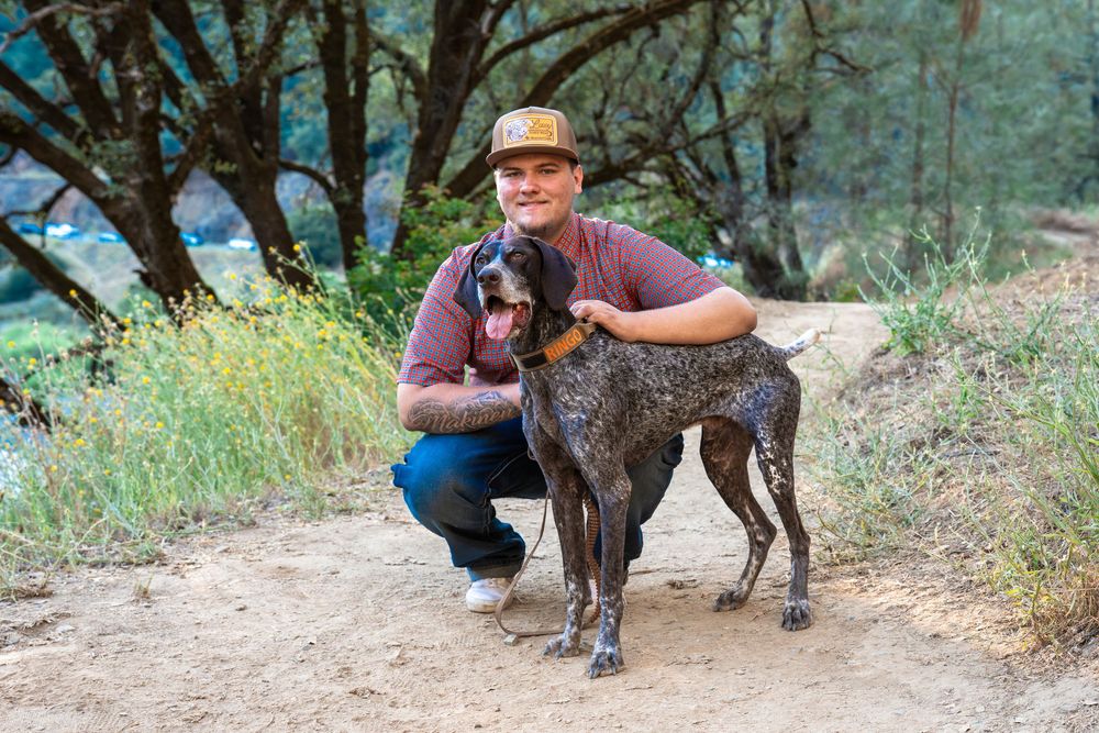 Man in cap kneeling beside a German Shorthaired Pointer dog on a dirt trail surrounded by trees.