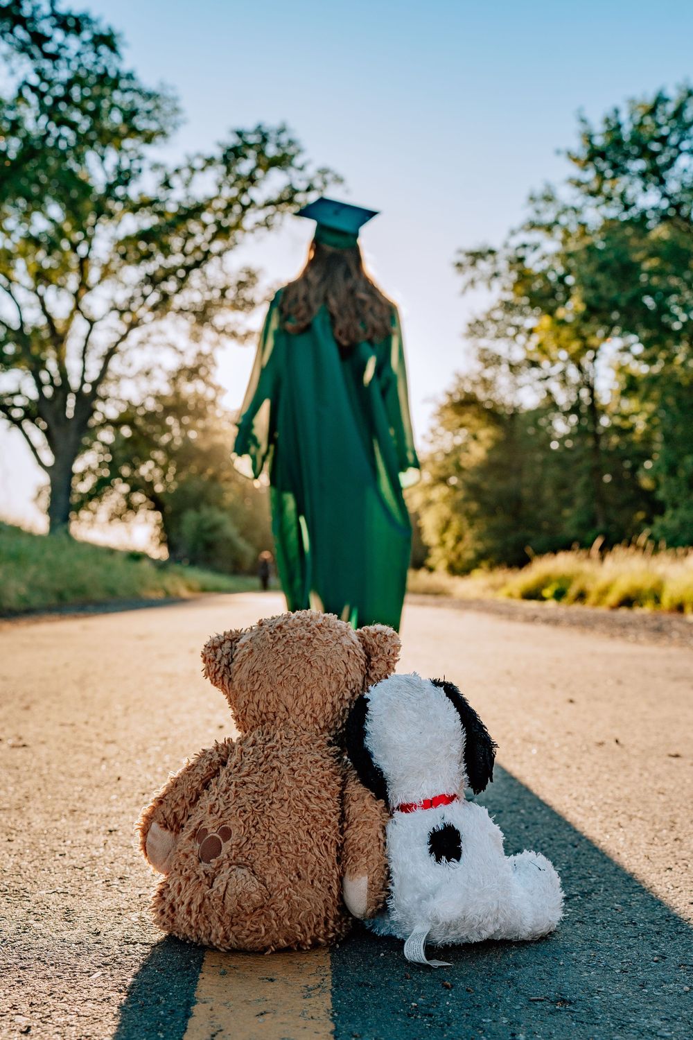 Graduate in green cap and gown walks away from two stuffed animals, a teddy bear and Snoopy, on a sunny path.