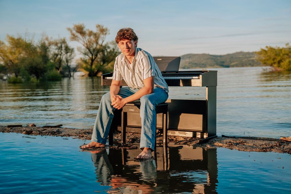 Young man sitting on a piano in shallow water with a scenic river and hills in the background.