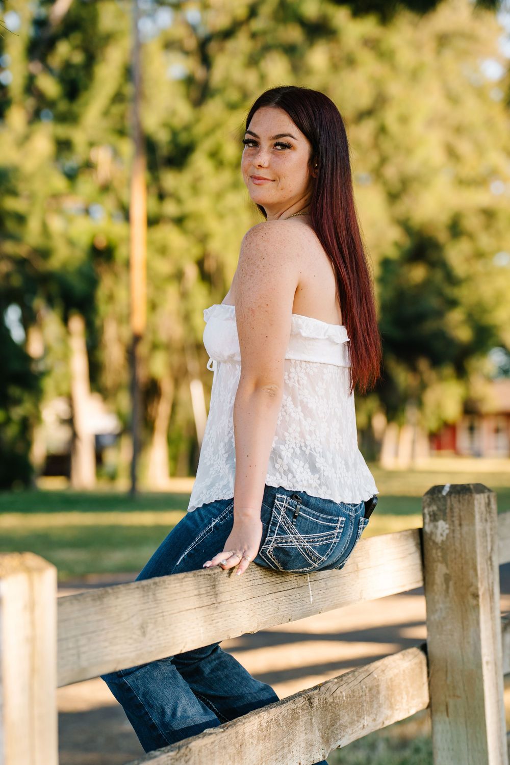 Young woman in white lace top and jeans leaning on wooden fence in sunny park setting.