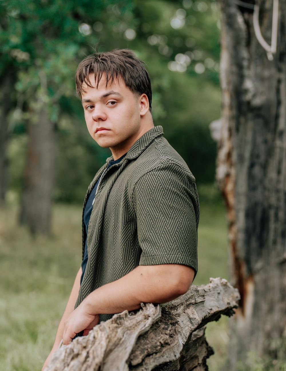 Young man in green shirt leaning on a weathered log in a lush outdoor setting, gazing thoughtfully.