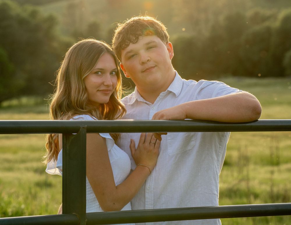 Young couple posing together at golden hour by a farm fence, surrounded by green fields.