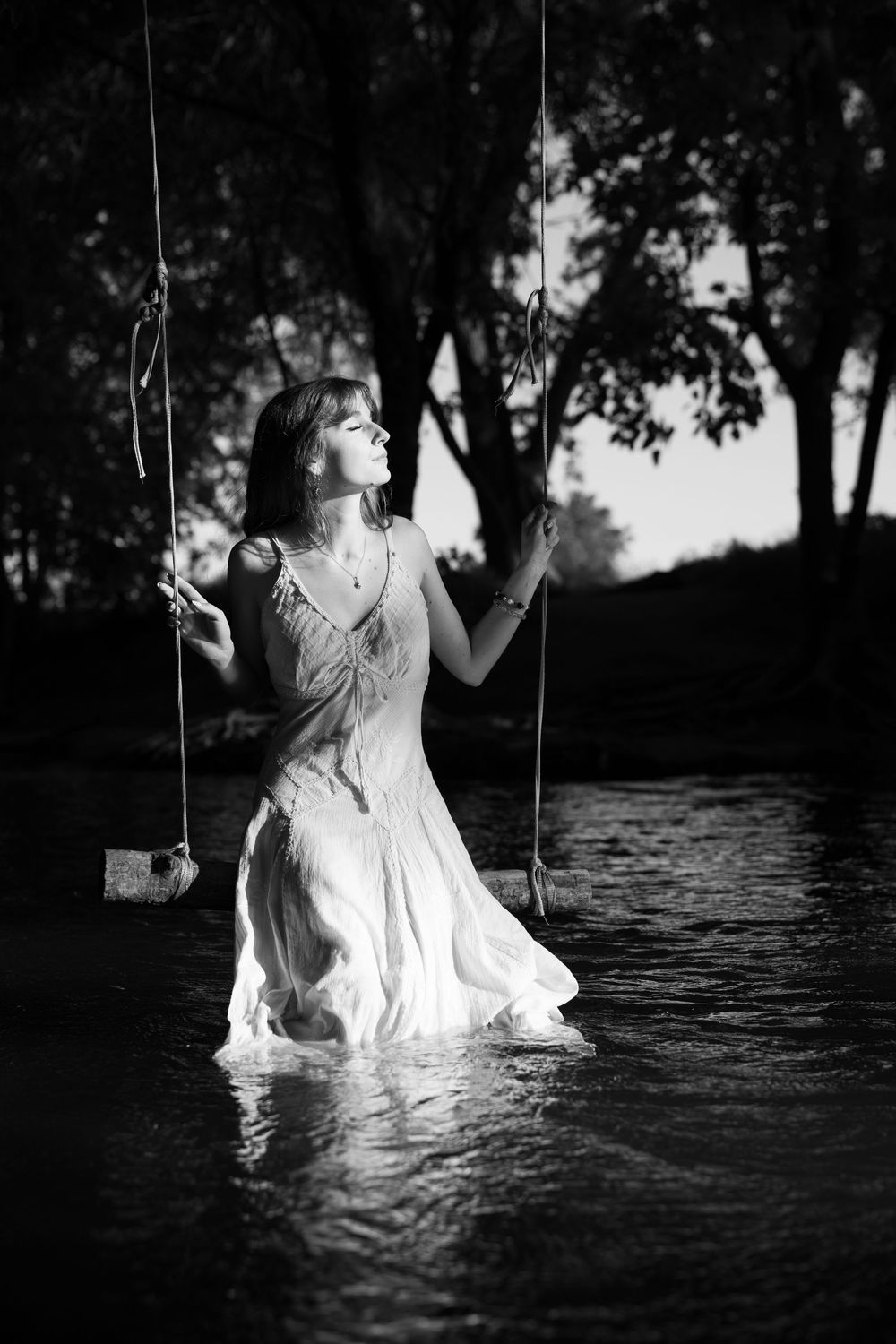 Black and white photo of a woman in a white dress standing in a river, holding swing ropes among trees.