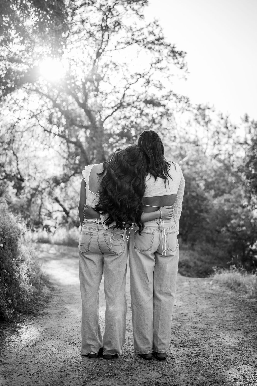 Two women with long hair embrace on a sunlit forest path, wearing matching jeans, photographed in black and white.