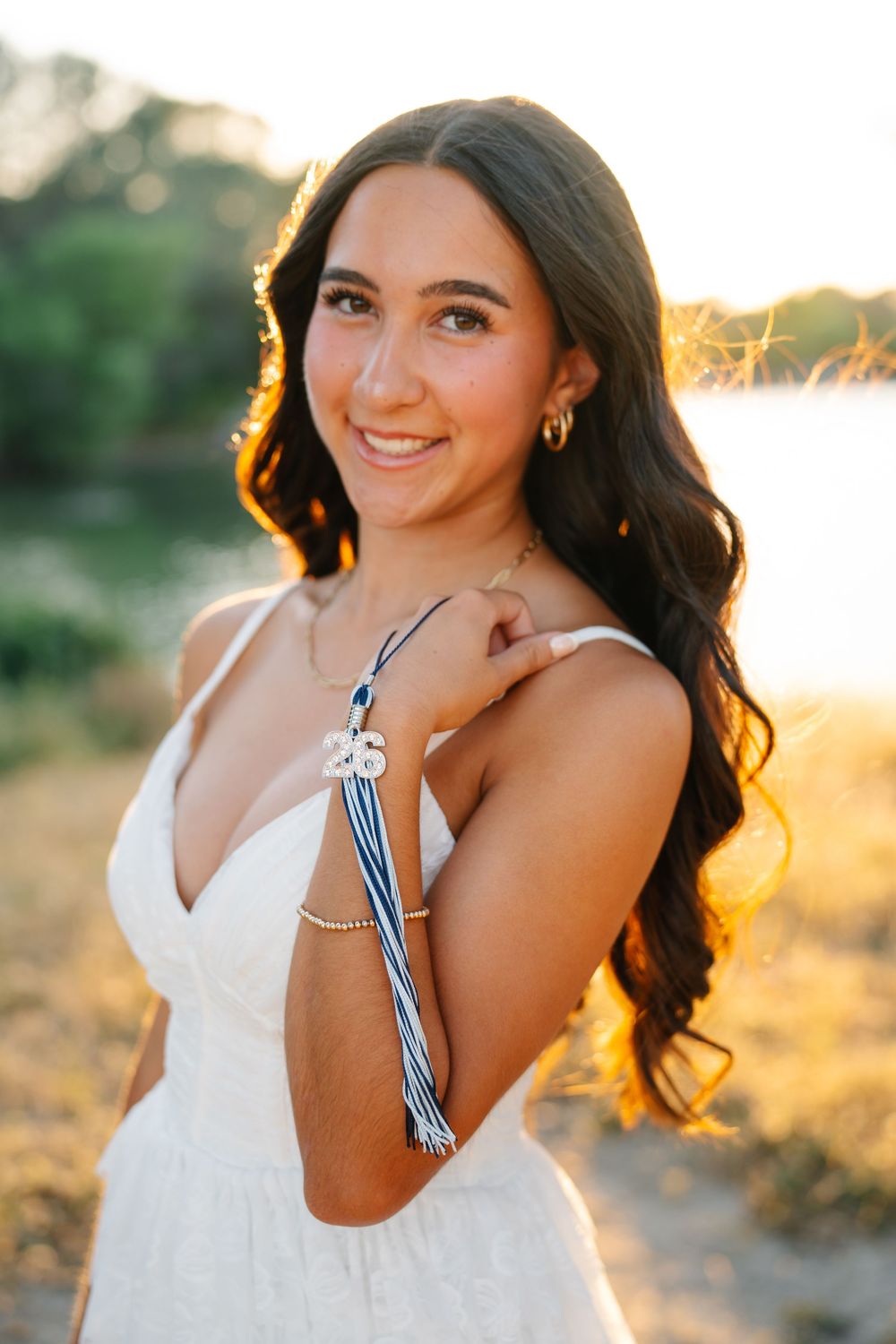 Smiling young woman in white dress holding graduation tassel outdoors at golden hour.