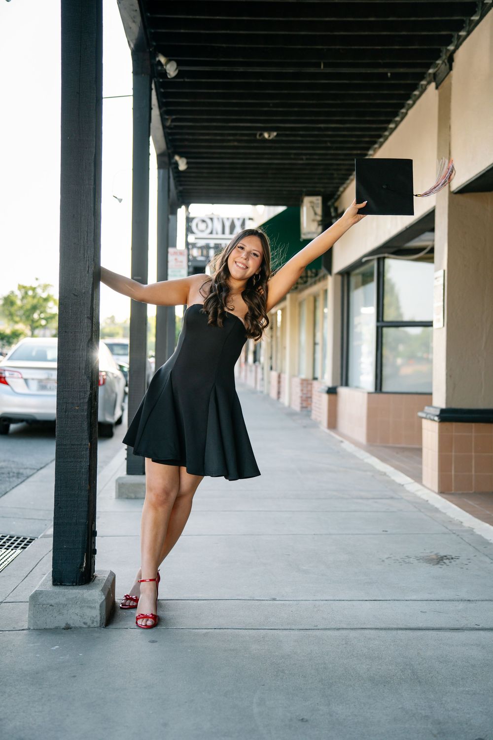 Young woman in black dress and red heels posing playfully on urban sidewalk under storefront awning.