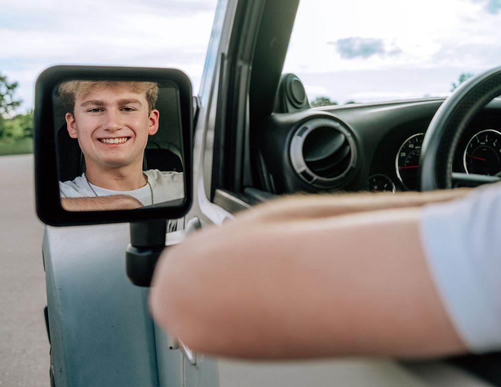 Smiling teenage boy reflected in a car side mirror while sitting in the driver's seat of a vehicle.