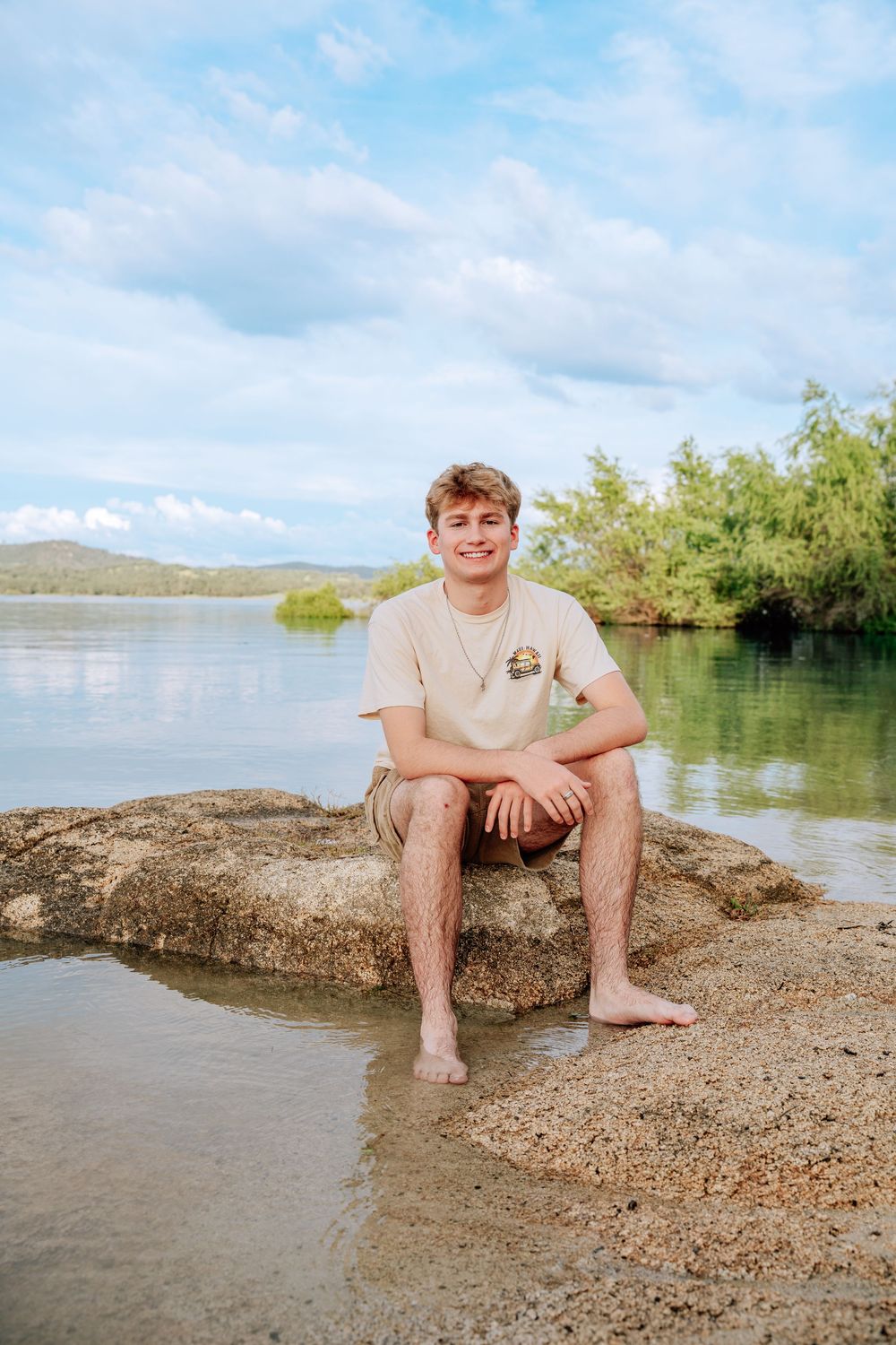 Young man sitting barefoot on a rock by a calm lake, smiling, wearing a beige t-shirt and shorts on a sunny day.