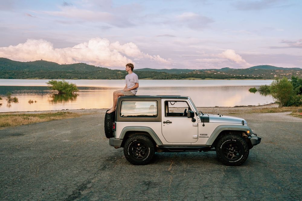 Man sitting on top of a white Jeep Wrangler parked by a scenic lake with mountains at sunset.