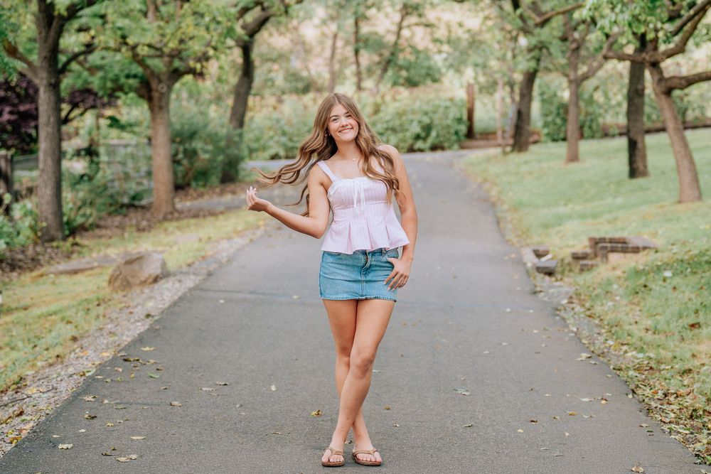 Young woman in denim skirt and white top posing on a tree-lined pathway outdoors on a sunny day.