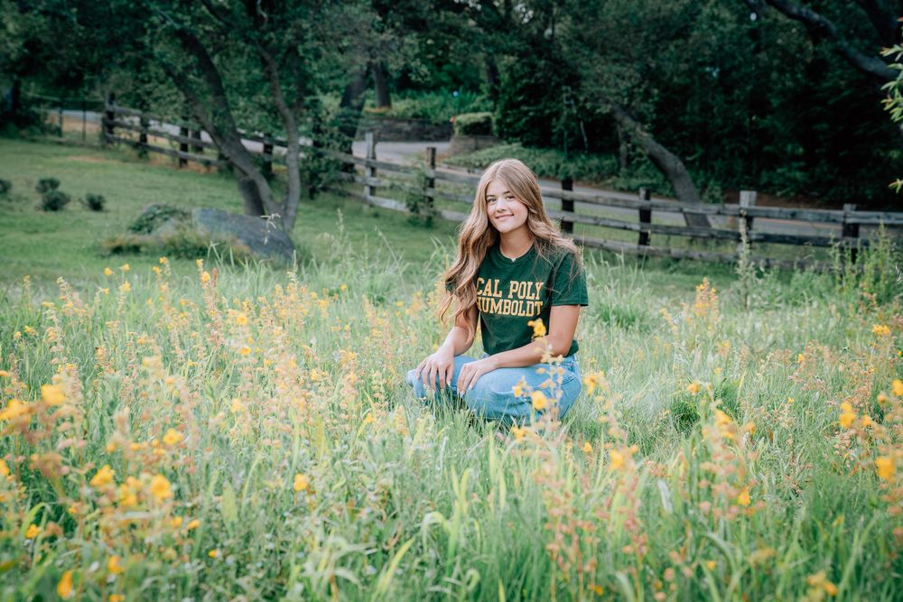 Young woman in green Cal Poly shirt sits smiling among wildflowers in a lush meadow with wooden fence backdrop.