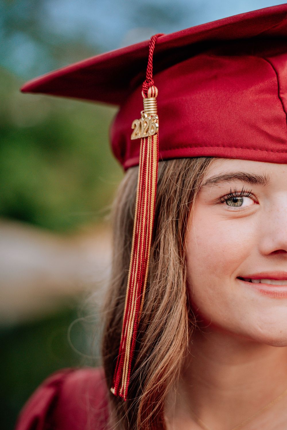 Smiling graduate wearing a red cap and tassel with 2023 charm, posing outdoors for a senior portrait photo.