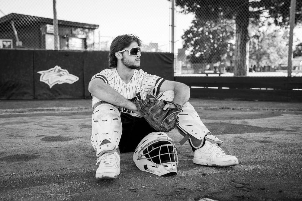 Baseball catcher in uniform sits on field ground with helmet and glove, wearing sunglasses in black and white photo.