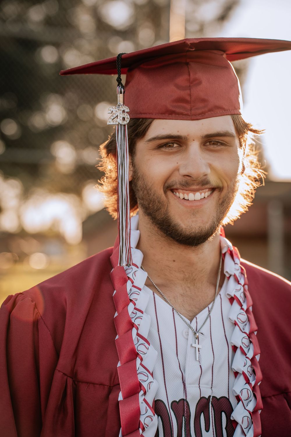 Smiling young man in red graduation cap and gown wearing a cross necklace, posing outdoors in warm sunlight.