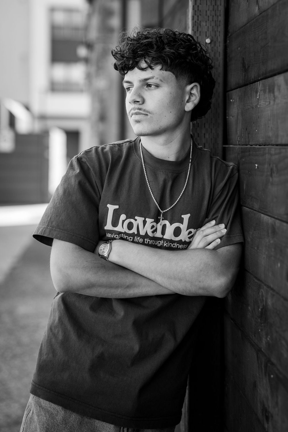 Young man with curly hair in a Lavender t-shirt leans against a wooden wall, arms crossed, in a black and white photo.
