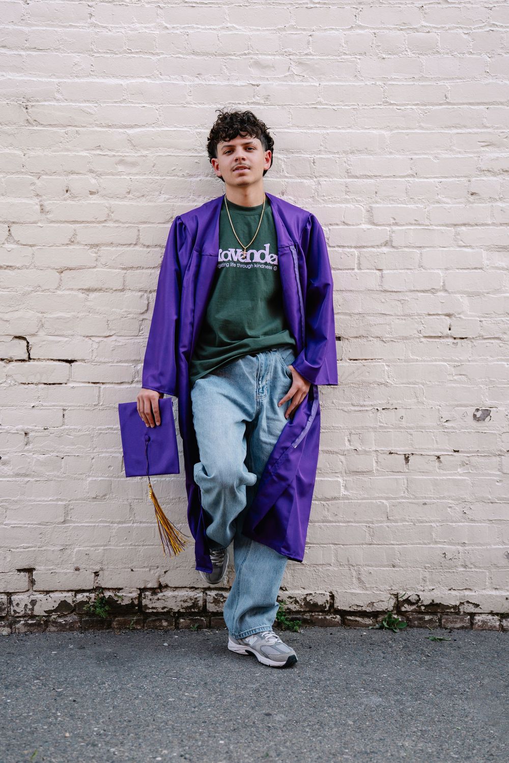 Young graduate in purple cap and gown leaning against white brick wall, wearing jeans and sneakers.