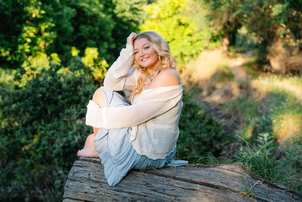 Smiling blonde woman sitting on a log in a sunlit forest, wearing a white sweater and light gray pants.