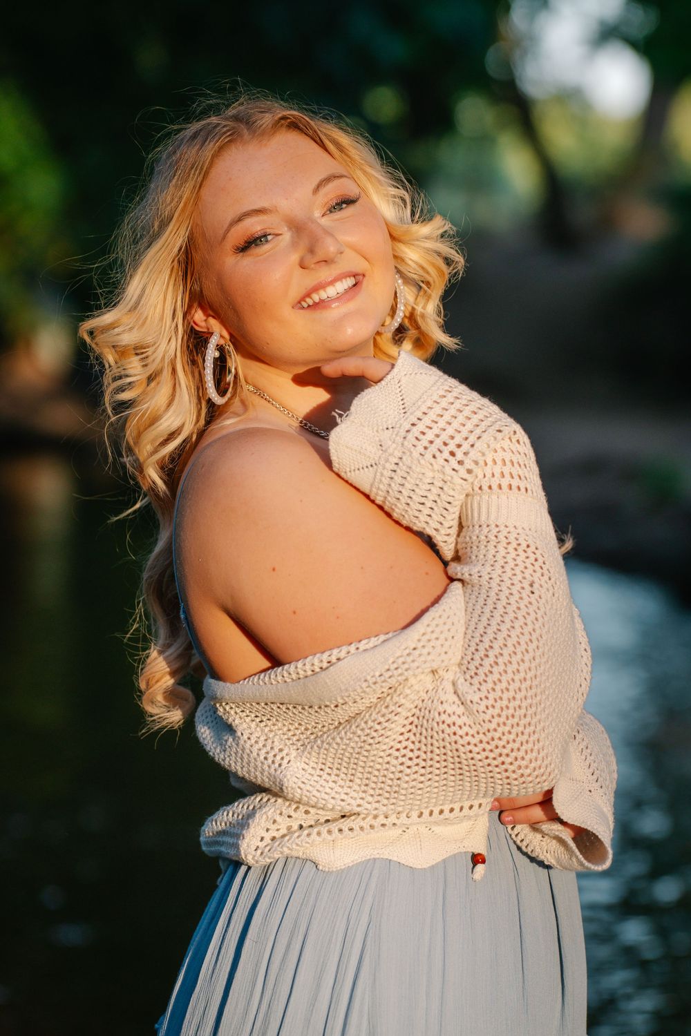 Smiling blonde woman in cream knit sweater and blue skirt poses outdoors near a sunlit creek.