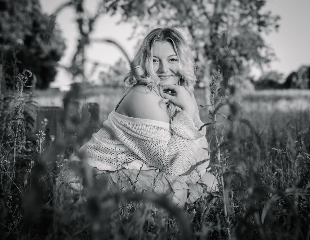 Black and white portrait of a smiling blonde woman in an off-shoulder top sitting among tall grass in a field.