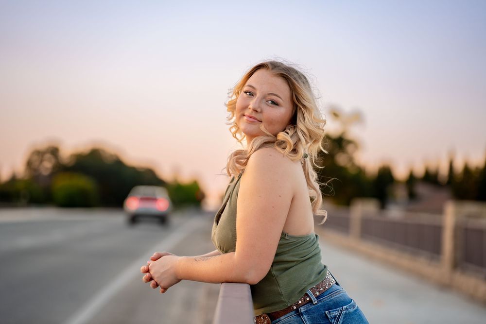 Young blonde woman in green top and jeans posing on a bridge at sunset with bokeh background.