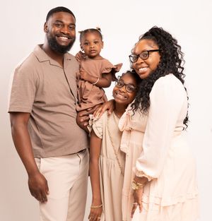 Family wearing coordinated beige and brown outfits pose together against white background for portrait session.