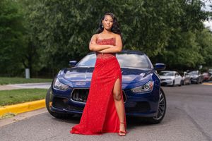 A person in a vibrant red formal dress poses elegantly next to a blue luxury car in an outdoor setting.
