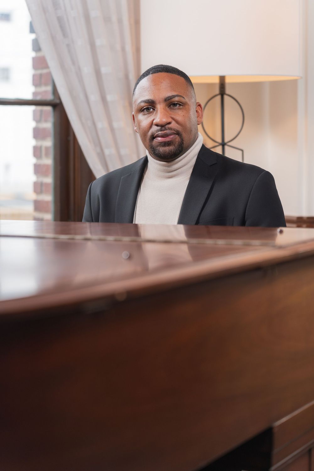 A person in a dark suit sits at a wooden piano in an elegant indoor setting.