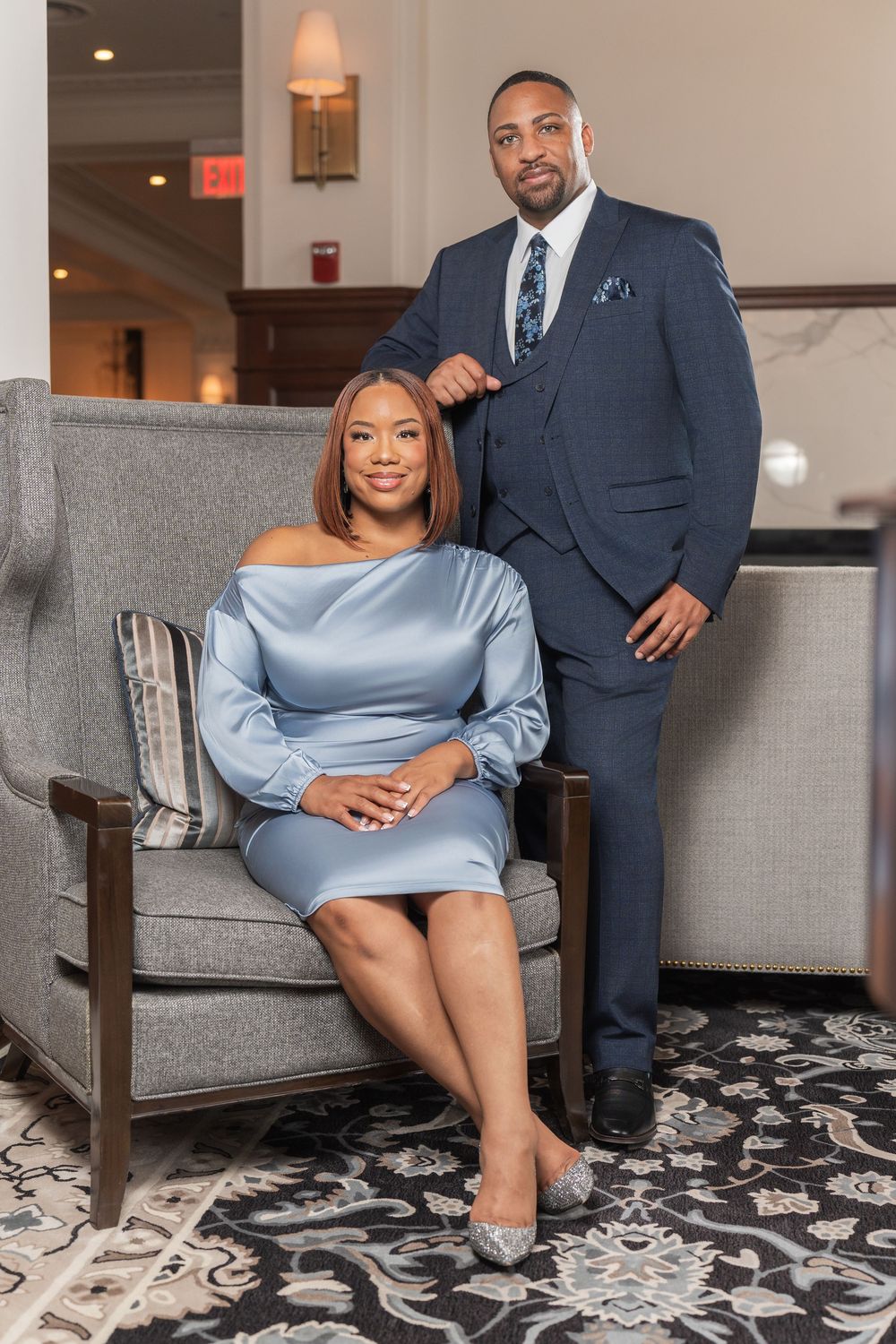 A couple poses on a grey couch, with one person wearing a light blue dress and another in a navy suit.