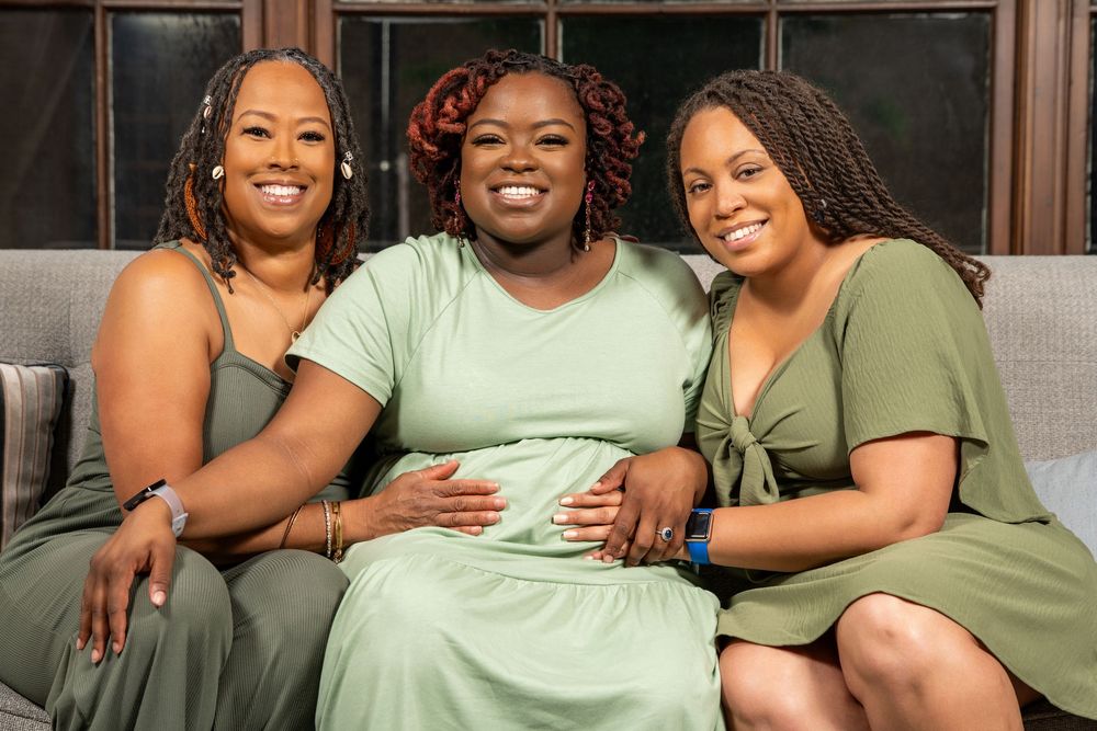 Three women wearing green dresses smile while sitting together on a grey couch.