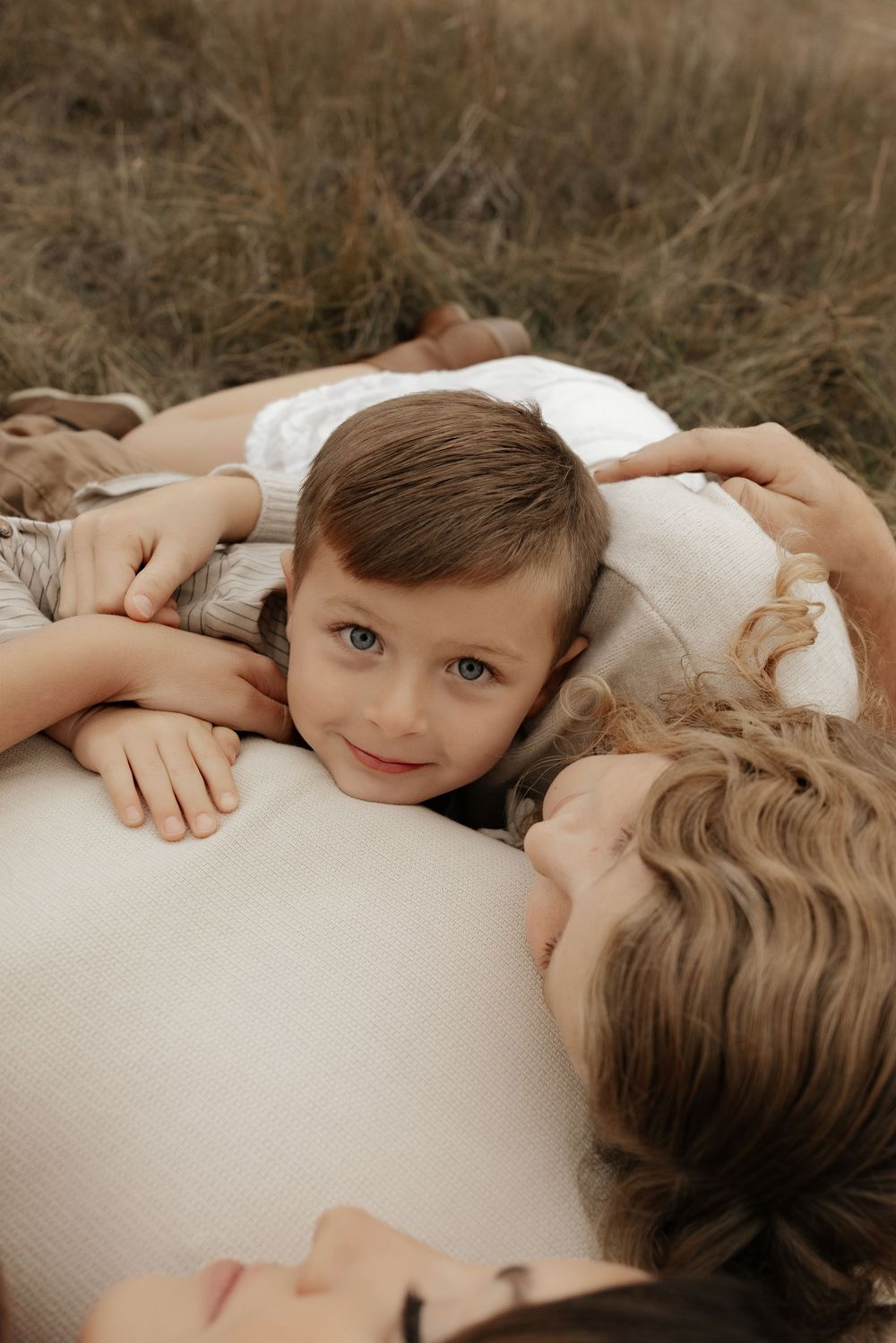 A young boy with blue eyes rests his head on a pregnant woman's belly while lying in a grassy field.