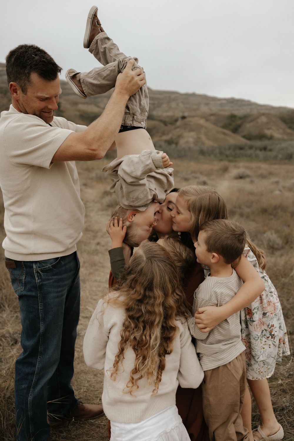 A joyful family shares kisses outdoors, with dad holding a child upside down against the alberta badlands during a photoshoot