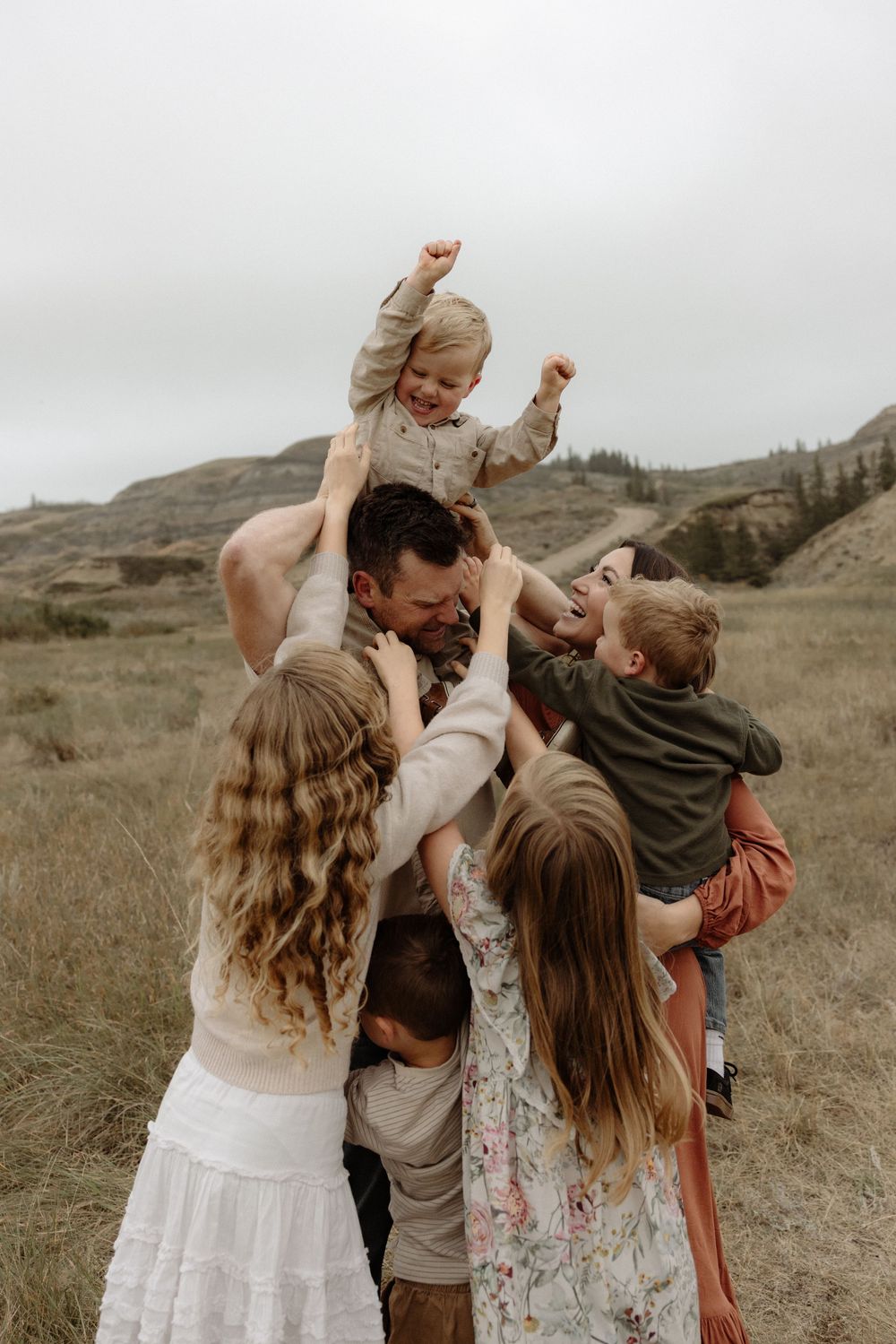 A joyful family group hug outdoors in a grassy field with rolling hills, children being lifted and embraced by parents.