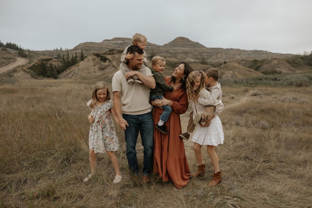Family of six posing together in a golden field with rolling hills, wearing warm earth-toned outfits for an outdoor portrait.