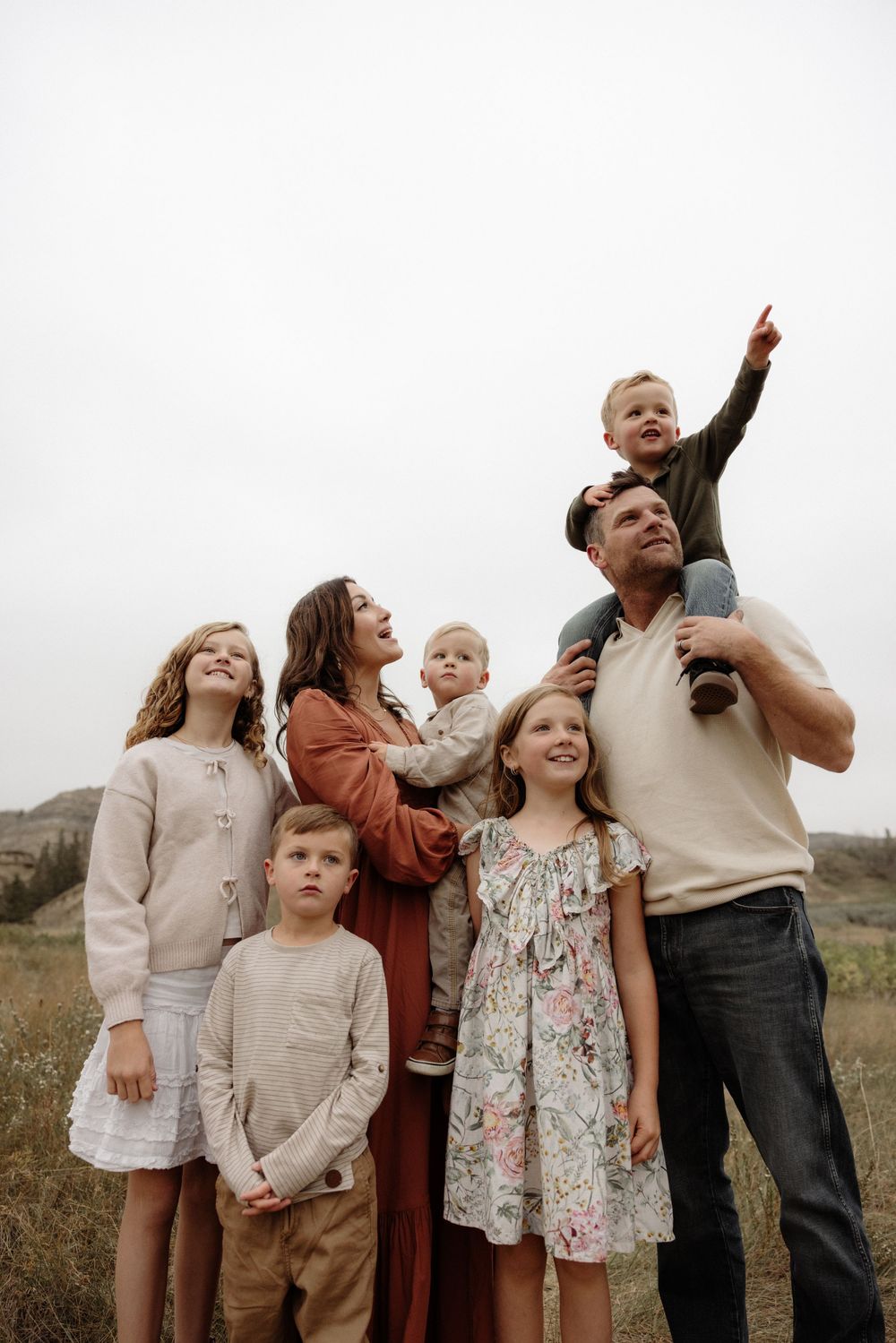 Family of seven poses outdoors in a field, parents with five children, one toddler pointing skyward.