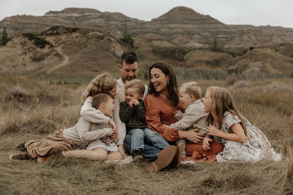 Family of six sitting together in a golden grass field with rocky hills, sharing a joyful moment outdoors.