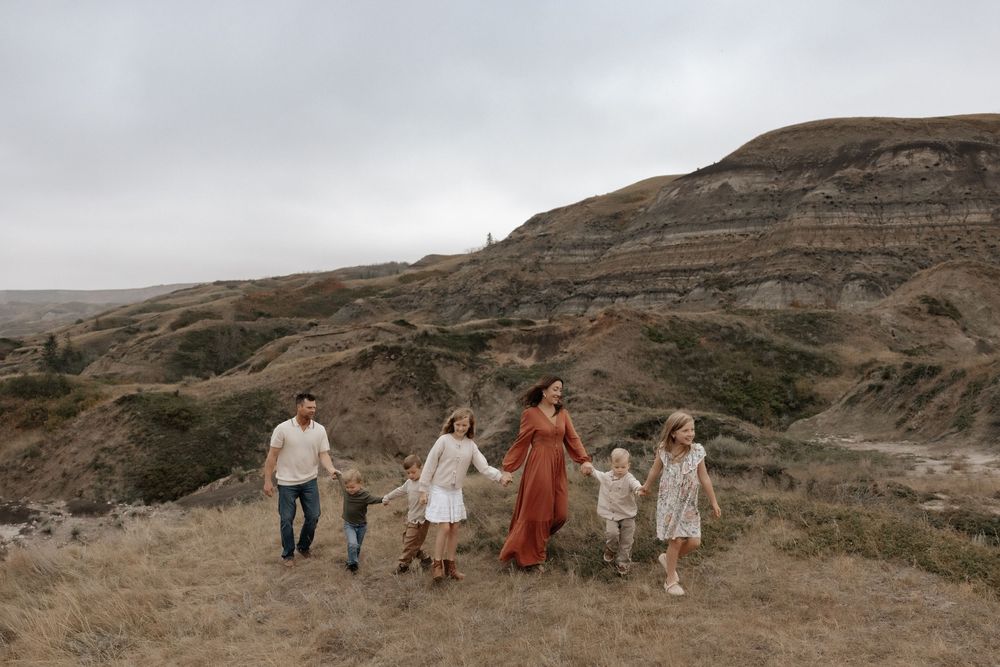 Family of six walks hand-in-hand through rugged badlands landscape under overcast skies.