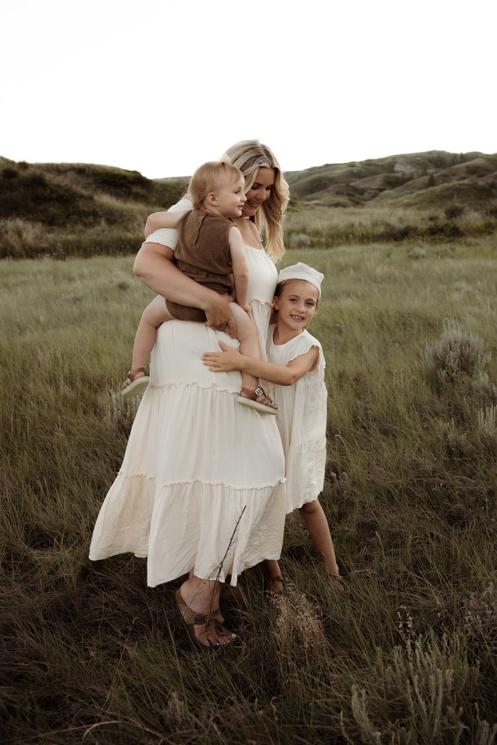 A mother in a white dress holds her toddler while her young daughter hugs her in a lush green meadow.
