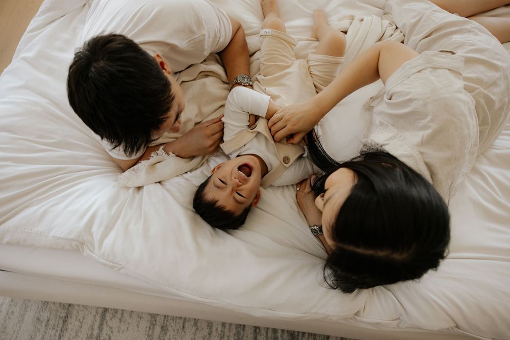 Two parents playfully laughing with their toddler on a white bed, enjoying a joyful family moment together.