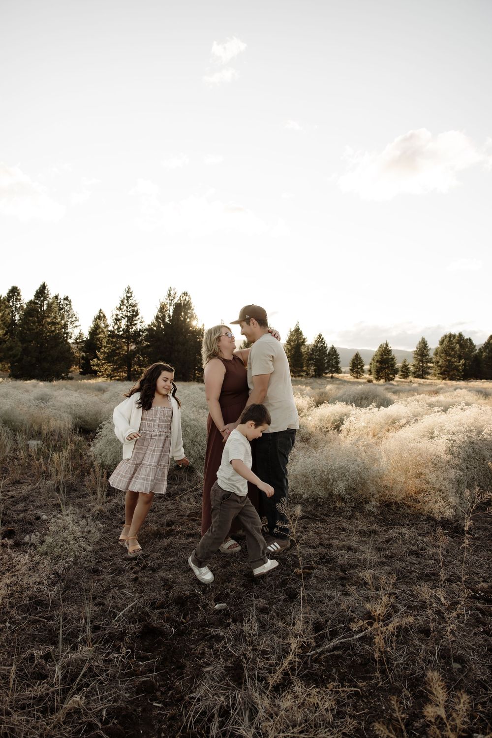 Family of four enjoying a golden hour stroll through a scenic meadow with evergreen trees in the background.