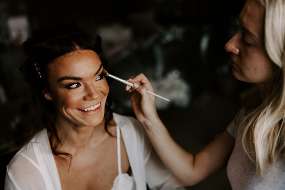 Makeup artist applies finishing touches on bride during wedding day preparation in dark moody setting.